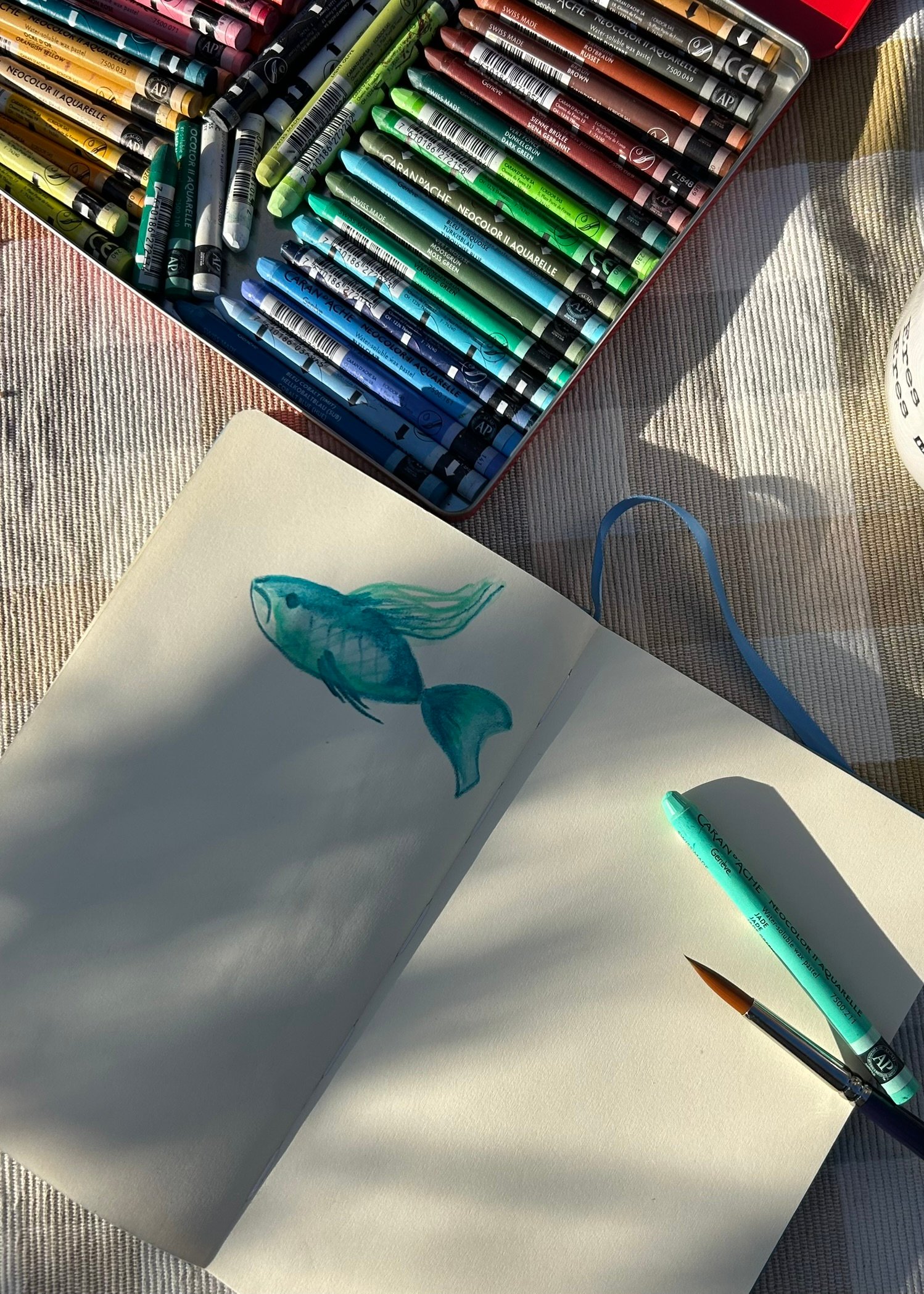 caja de ceras acuarelables de colores junto a un cuaderno con un dibujo de un pez