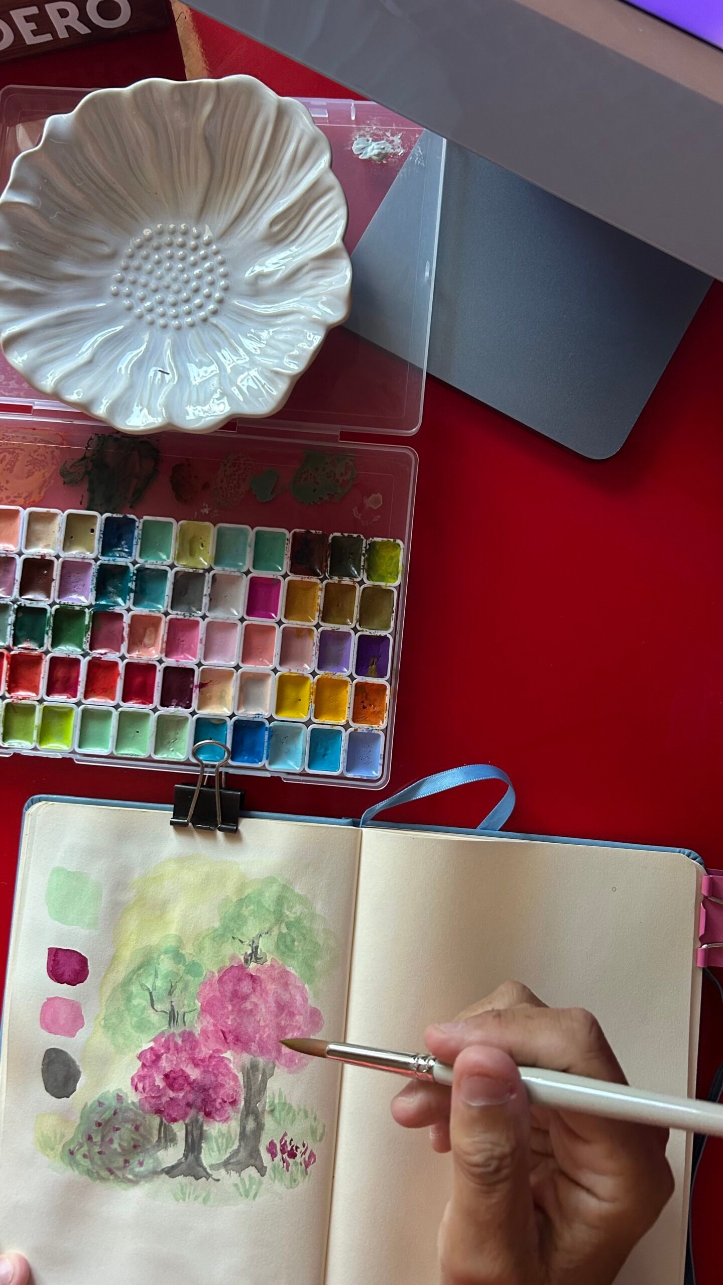 paleta de acuarelas de colores abierta sobre un paño rojo junto a un cuaderno