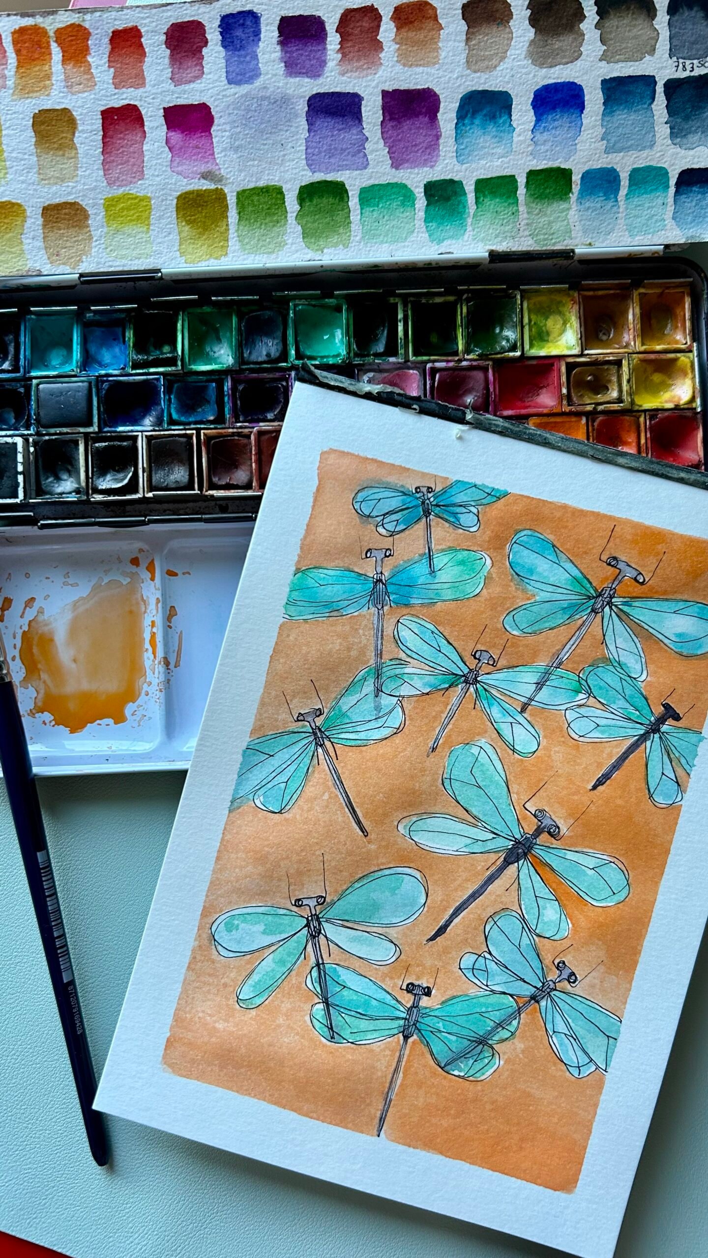 dibujo de libélulas azules sobre fondo naranja pintado en acuarela junto a una paleta de colores